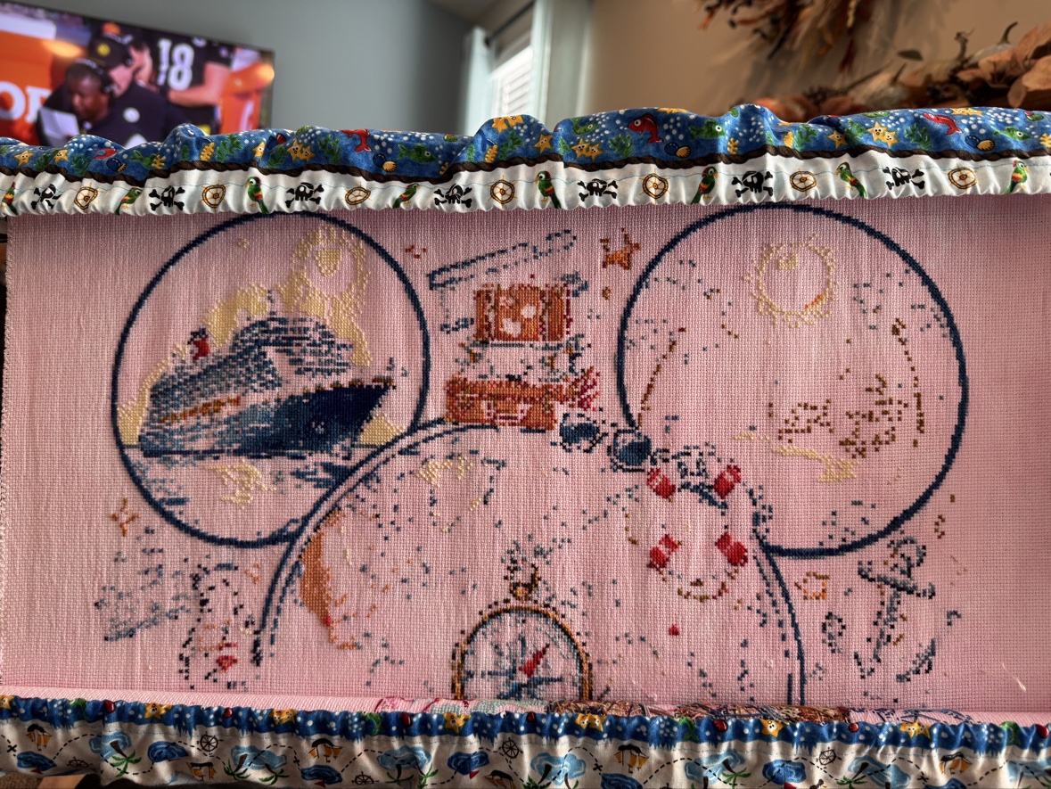 Anne Conrad&rsquo;s &ldquo;Magical Cruise&rdquo; cross stitch in progress on pale pink fabric in a scroll frame with pirate-print guards — the cruise ship stitched in the left ear, suitcases stacked at the top, and the compass rose taking shape below