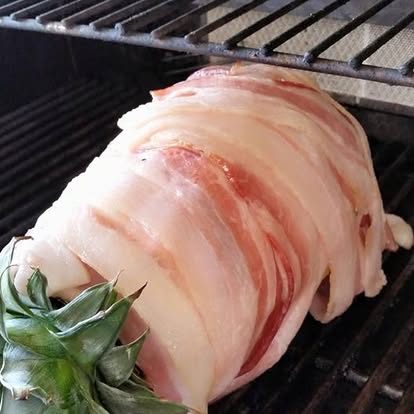 The Swineapple just on the grill — a pineapple wrapped in strips of raw bacon with its green crown back on, resting on the grill grates
